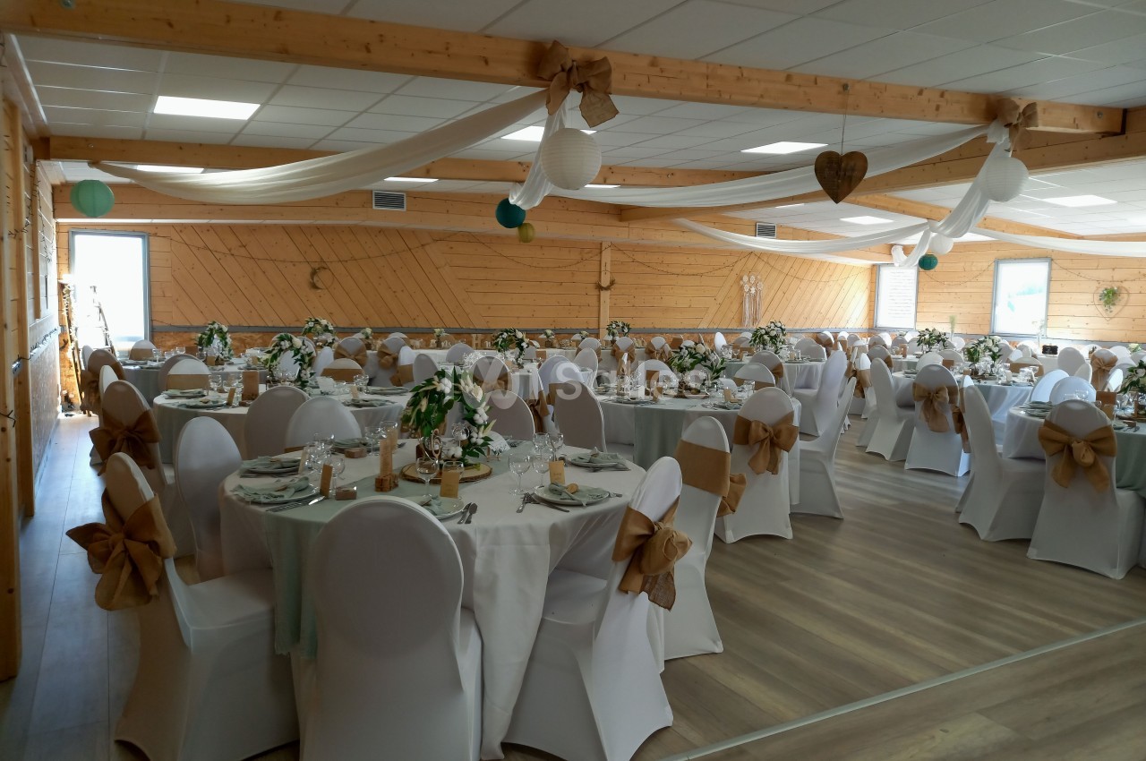 Salle de réception décorée avec des tables rondes, nappes blanches, chaises ornées de nœuds beiges et guirlandes suspendues.