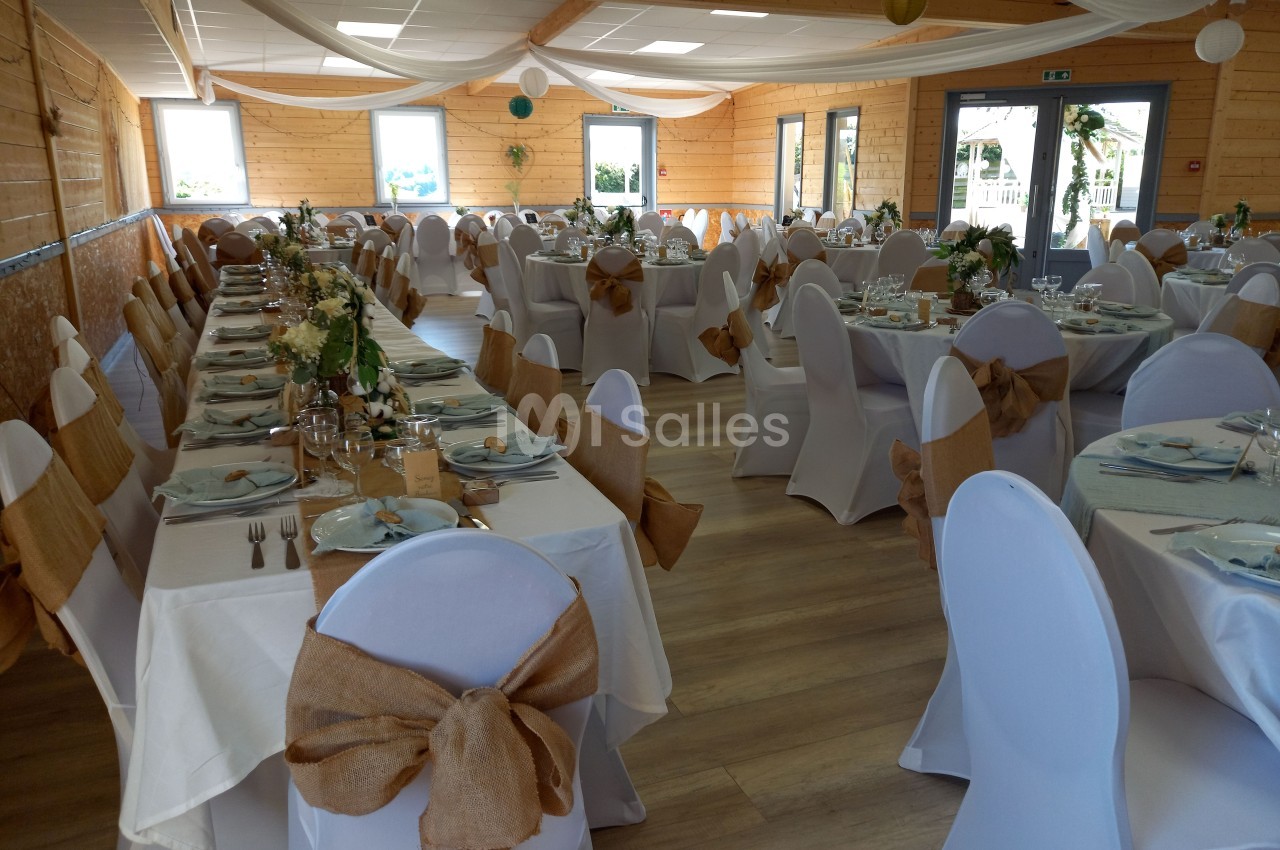 Salle de réception décorée pour un événement, avec tables dressées, nappes blanches et chaises ornées de nœuds en toile de…