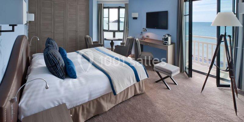 Chambre lumineuse avec lit double, bureau, fauteuils et balcon offrant une vue sur la mer.
