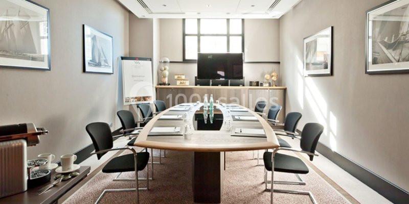 Salle de réunion lumineuse avec une grande table ovale, des chaises noires, un écran et un tableau de présentation.