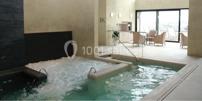 Piscine intérieure avec jets d'eau et bain à remous, entourée de chaises et baignée de lumière naturelle.