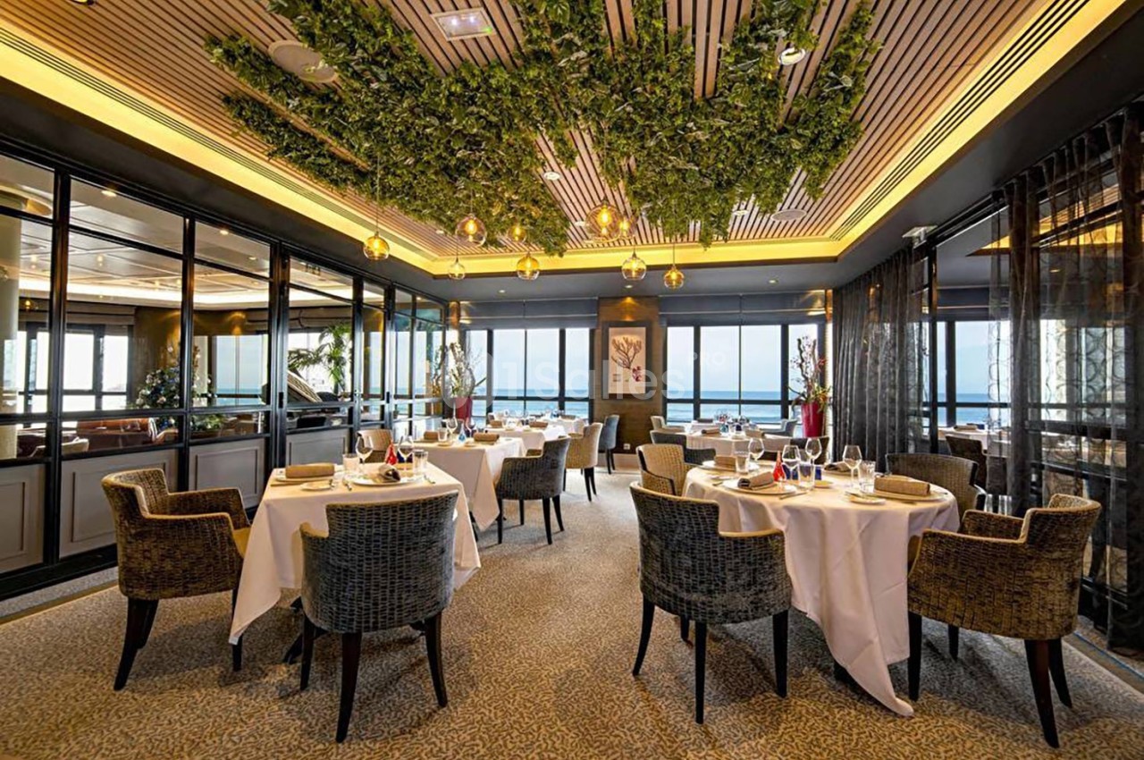 Salle de restaurant élégante avec tables dressées, chaises rembourrées, plafond végétalisé et vue sur l'extérieur.