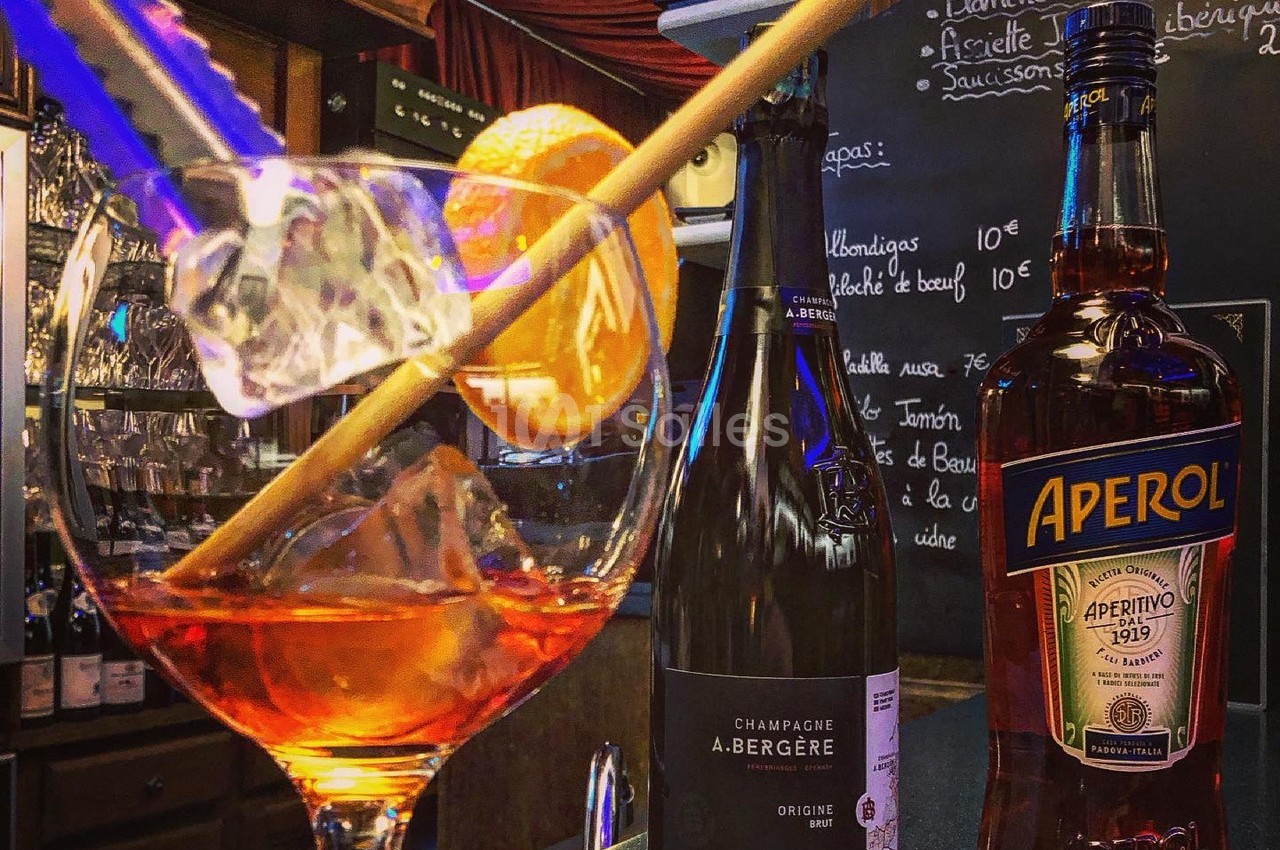 Verre de cocktail avec tranche d'orange et glaçons, accompagné de bouteilles de champagne et d'Aperol sur un bar.