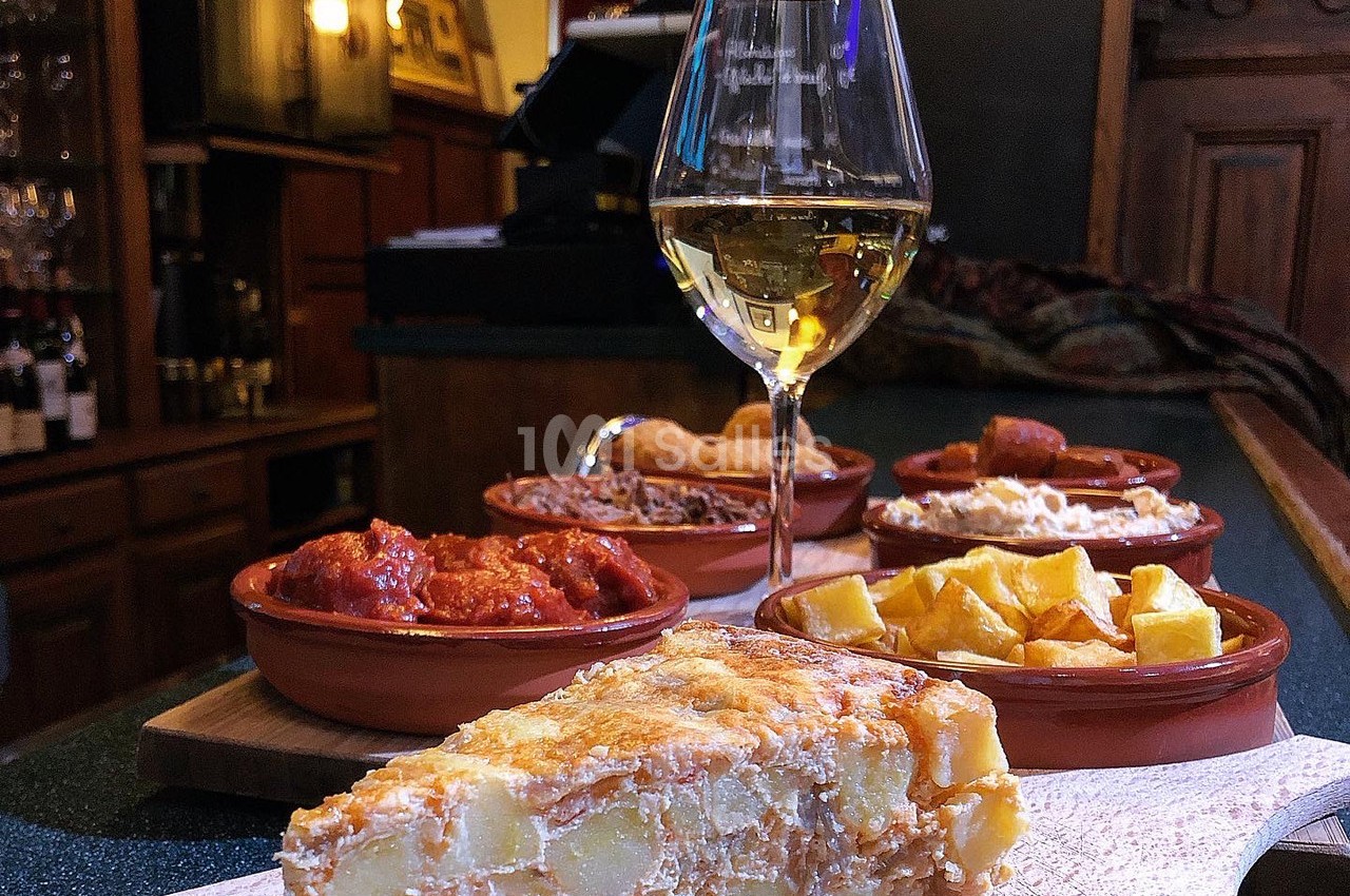 Assortiment de tapas espagnoles avec tortilla, patatas bravas et autres plats, accompagné d'un verre de vin blanc.