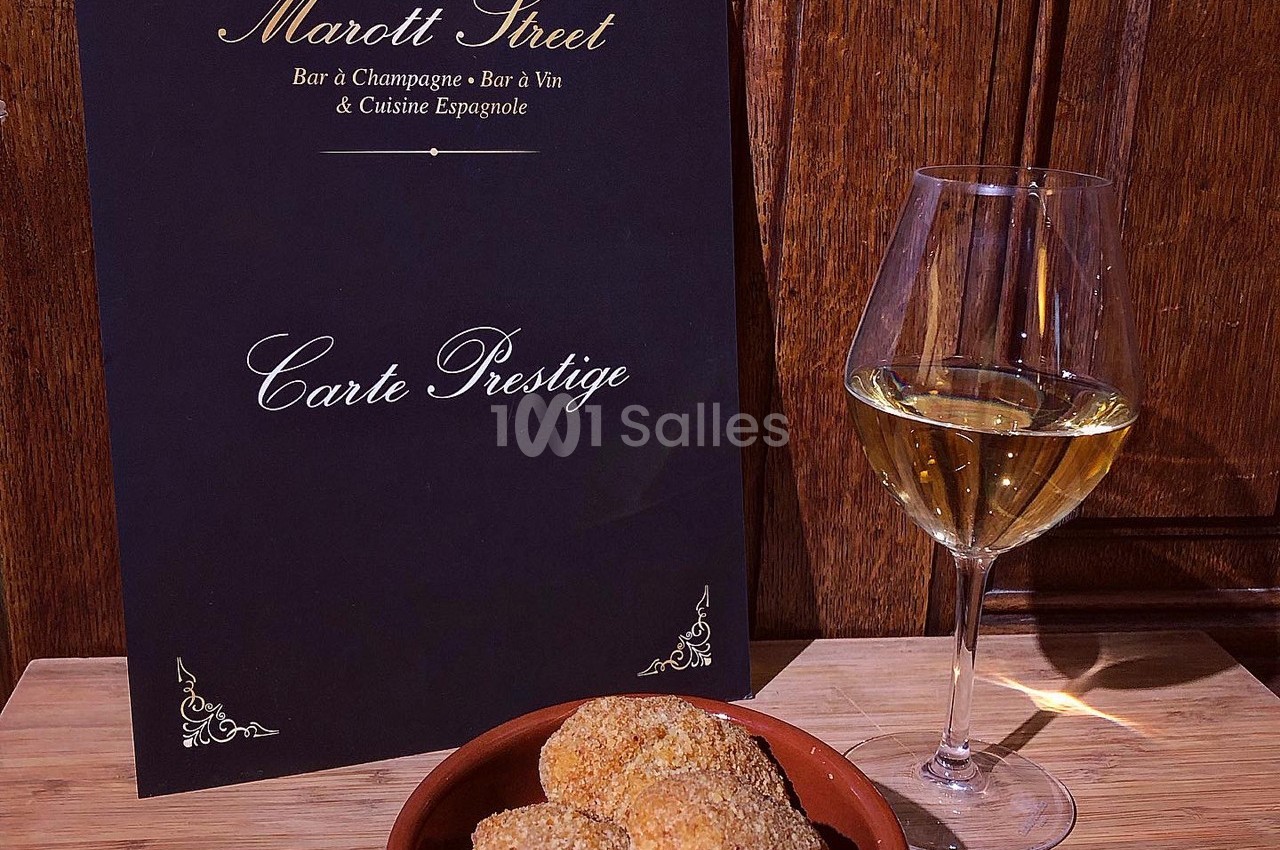 Carte de restaurant posée sur une table en bois avec un verre de vin blanc et une assiette de croquettes.