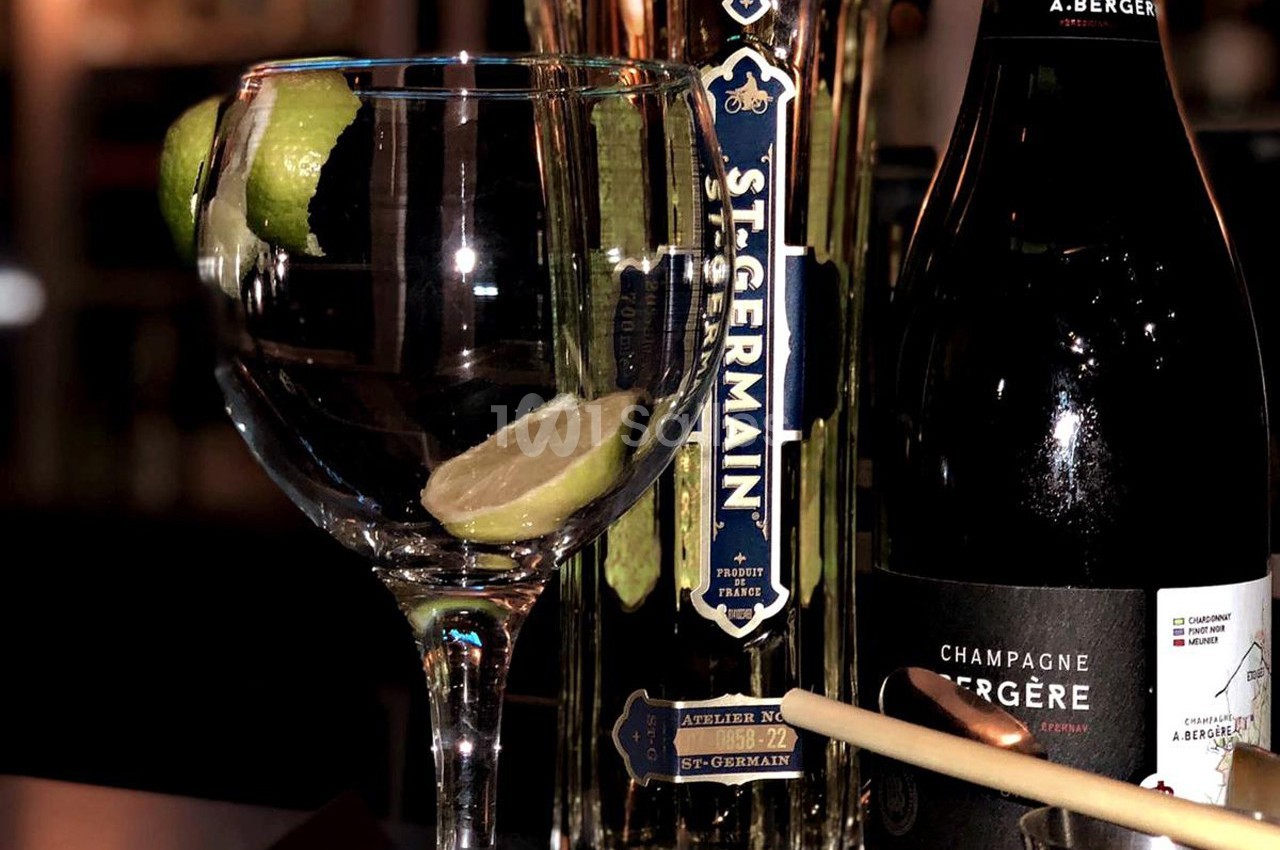 Un verre avec une tranche de citron vert, une bouteille de liqueur St-Germain et une bouteille de champagne A. Bergère.