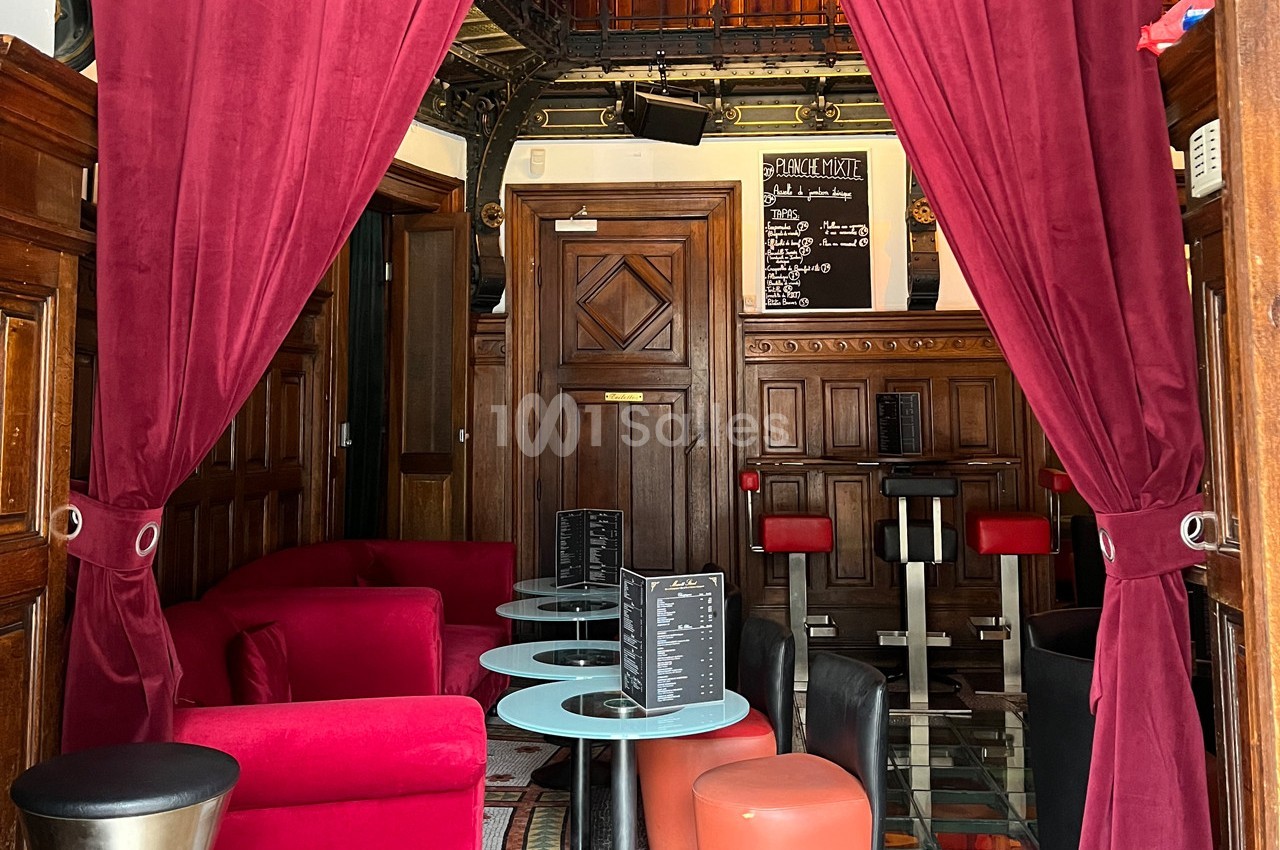 Espace intérieur avec fauteuils rouges, tables basses et rideaux ouverts, décoré de boiseries et d'un tableau noir au fond.