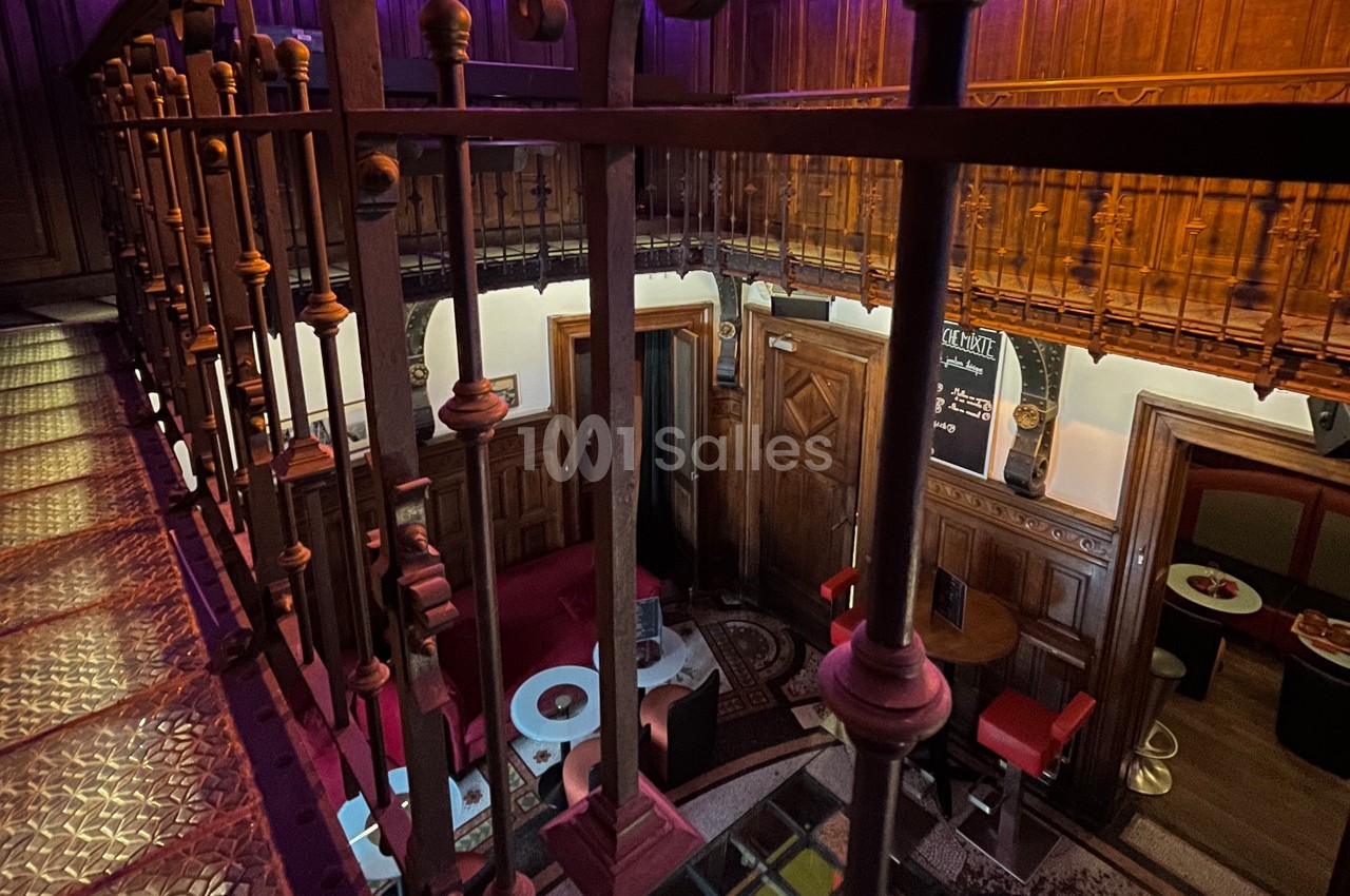 Vue intérieure d'un espace avec mezzanine en bois, balustrade métallique et coin salon avec tables et banquettes rouges.