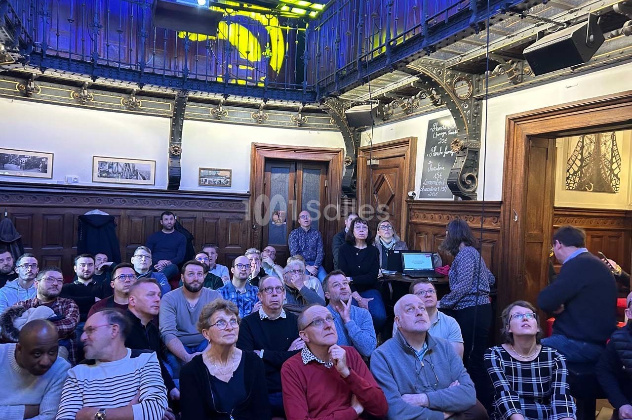 Un groupe de personnes assises dans une salle avec un décor en bois et une mezzanine métallique, regardant vers l'avant.