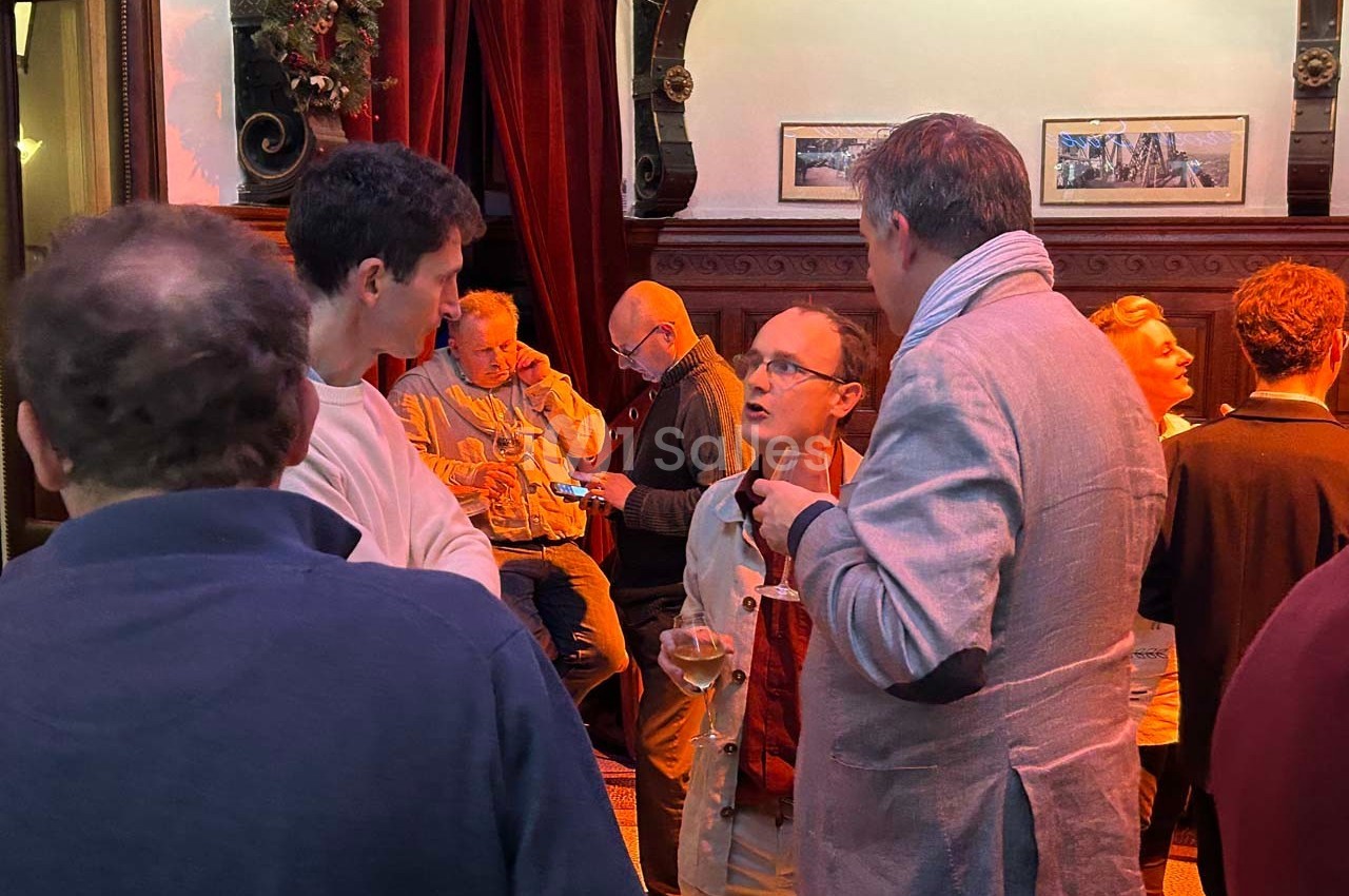 Des personnes discutent debout dans un intérieur chaleureux avec des rideaux rouges et des cadres accrochés au mur.