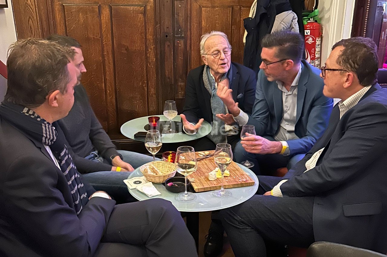 Cinq hommes discutent autour de petites tables avec des verres, des snacks et un décor intérieur en bois.