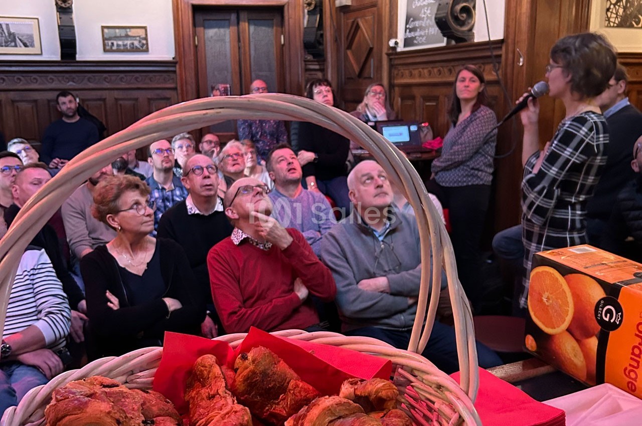Un groupe de personnes assises dans une salle en bois, écoutant une intervenante, avec des viennoiseries au premier plan.