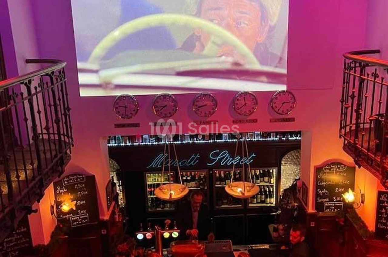 Vue intérieure d'un bar avec un écran projetant une scène de film, des horloges murales et un comptoir éclairé.