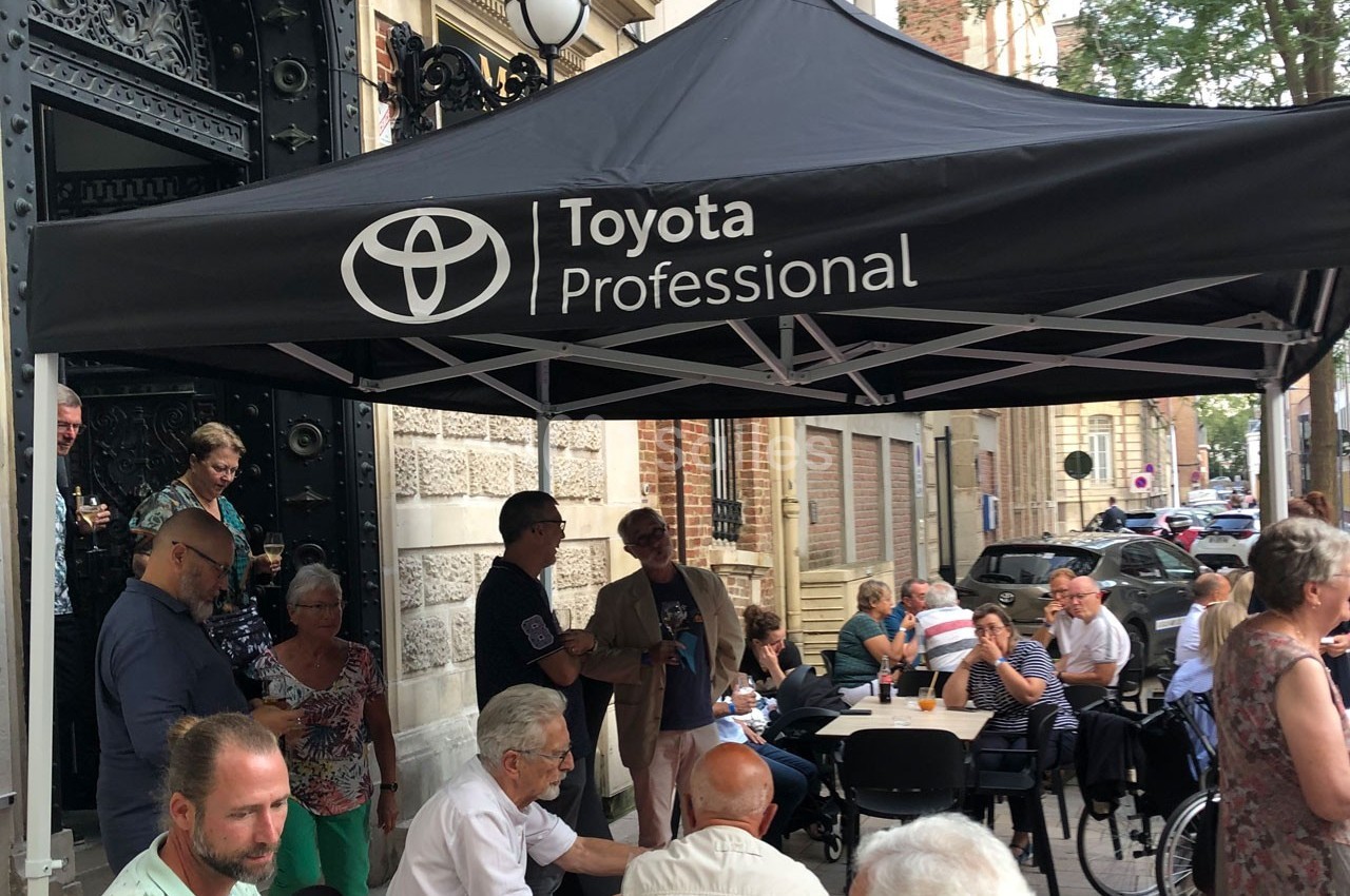 Groupe de personnes assises et debout sous un chapiteau noir marqué ’Toyota Professional’ dans une rue animée.