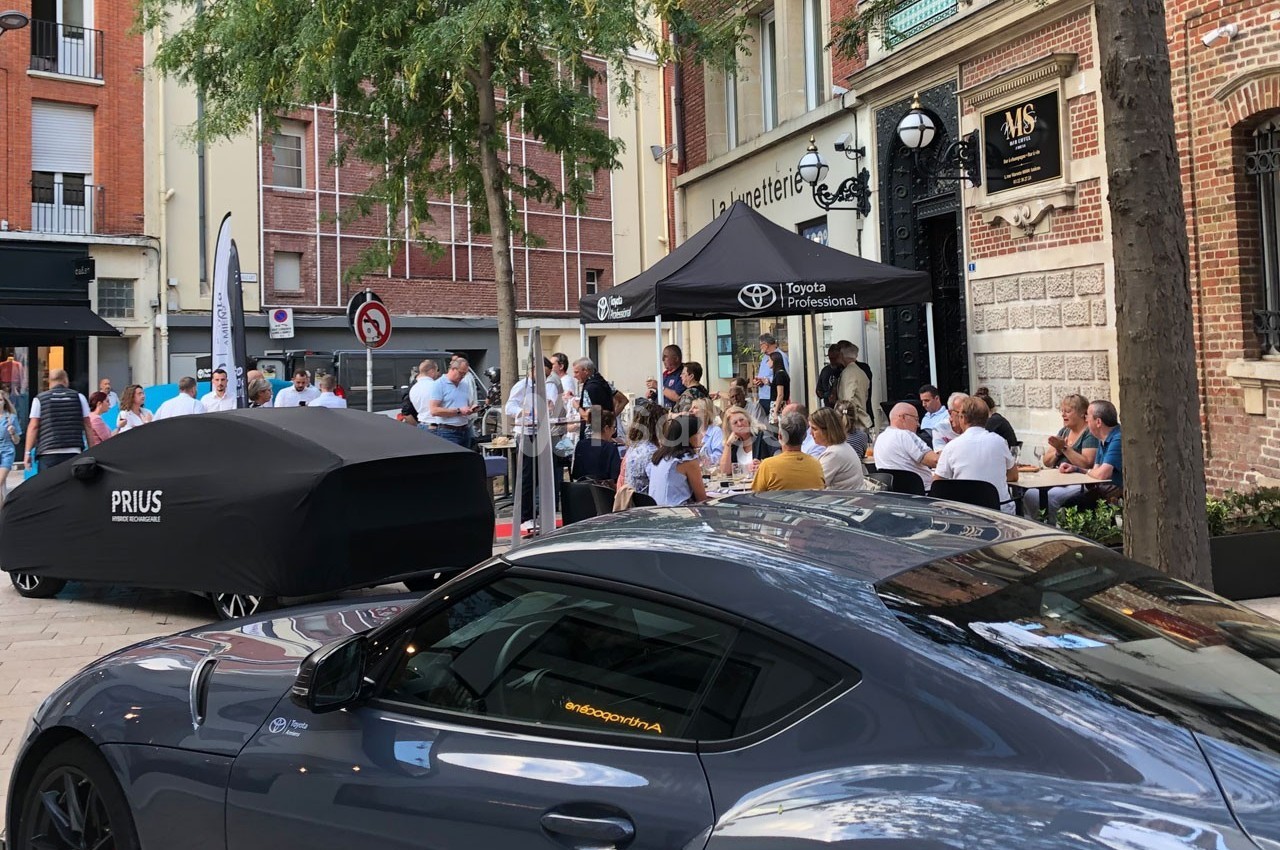 Voiture sportive grise stationnée devant un bâtiment, avec un événement rassemblant des personnes en arrière-plan.