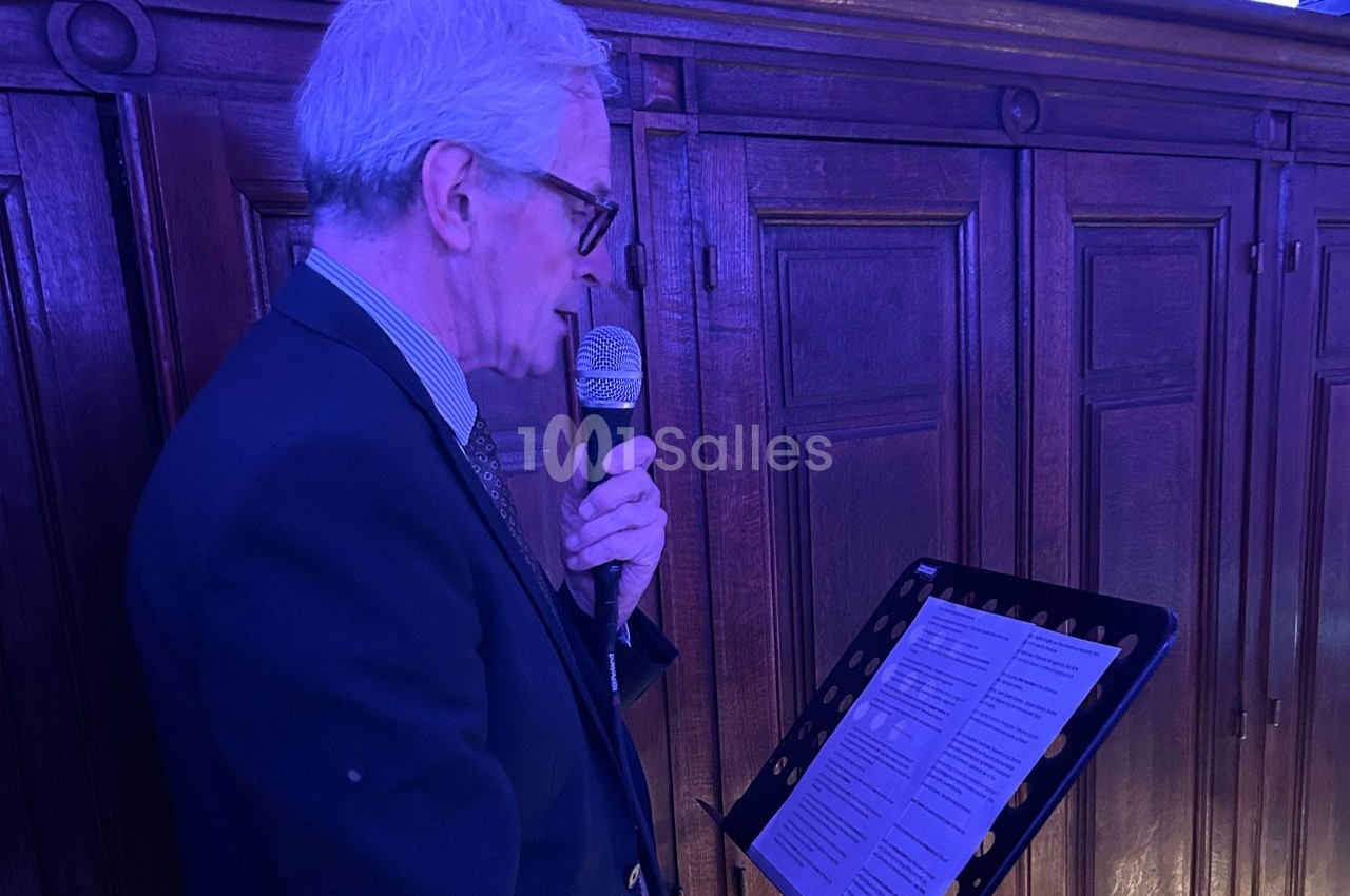 Un homme âgé en costume parle dans un micro, lisant un texte sur un lutrin, devant un fond en bois sombre.