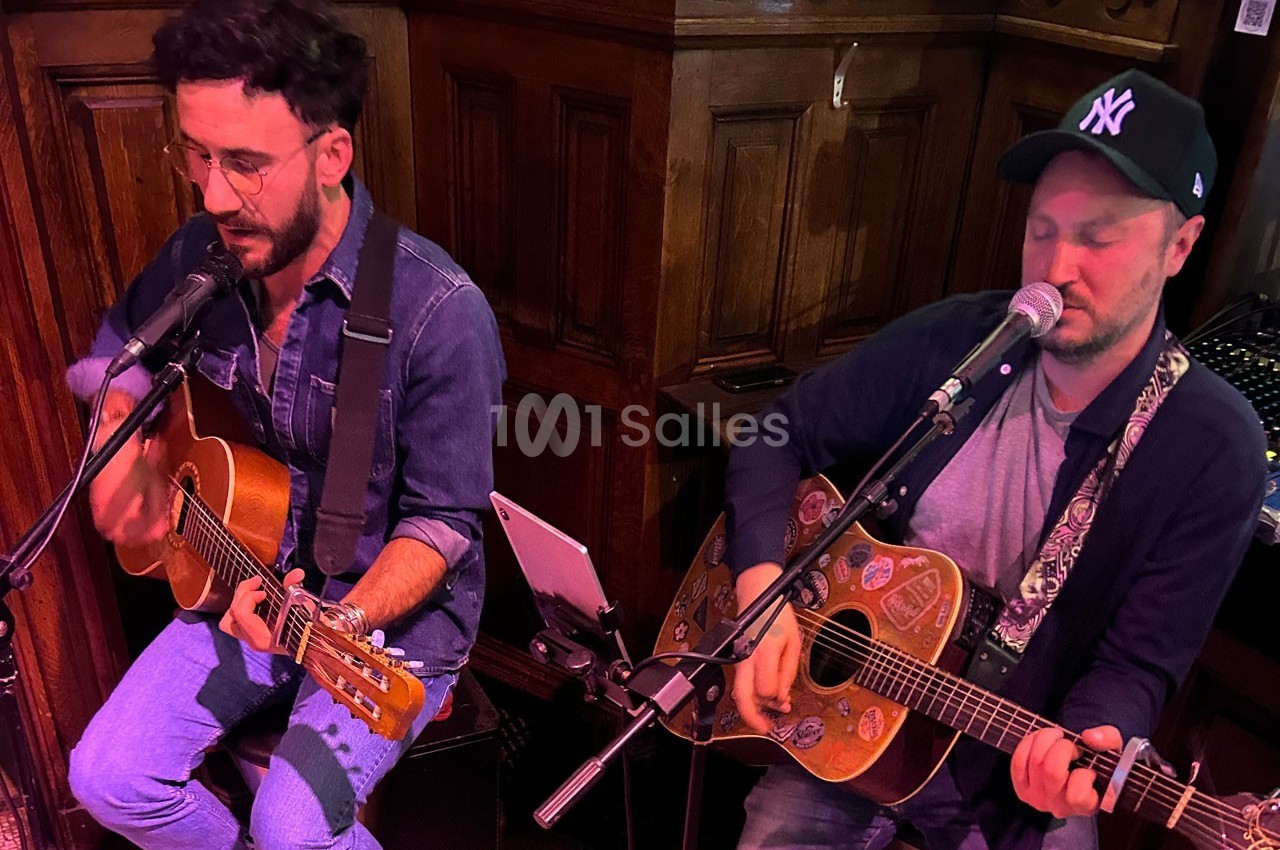 Deux musiciens jouent de la guitare et chantent dans un espace intérieur en bois, équipés de micros.