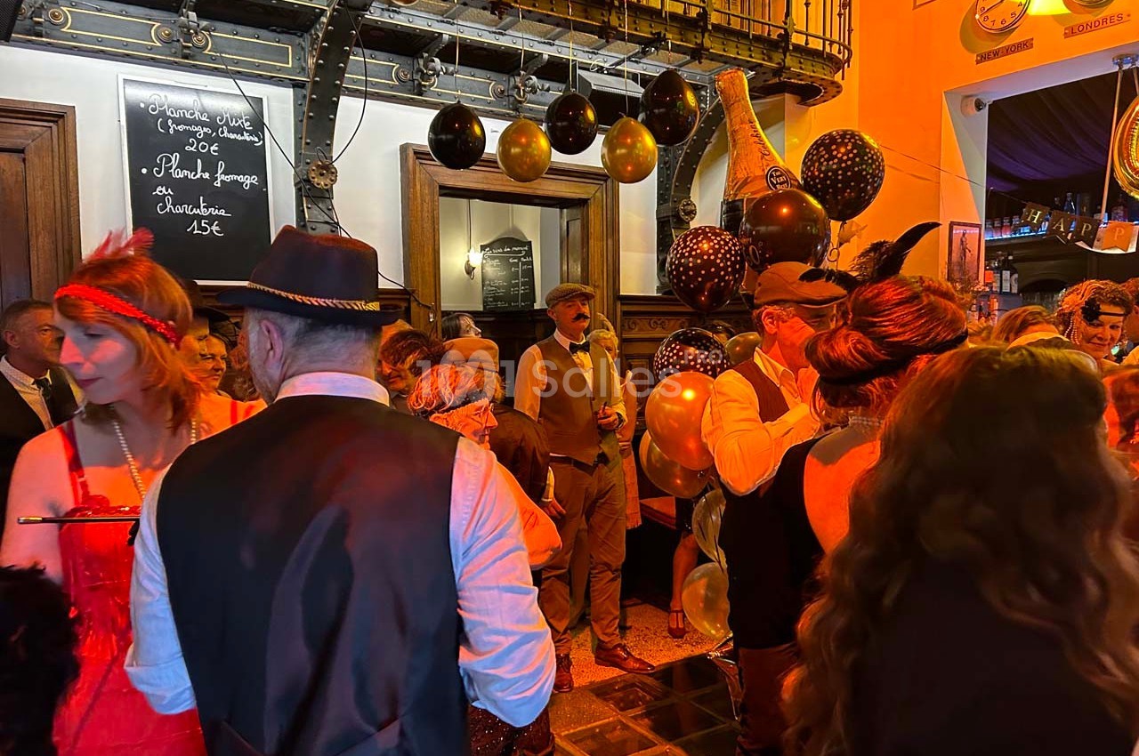 Personnes en tenue rétro rassemblées dans un bar décoré de ballons noirs et dorés, ambiance festive.
