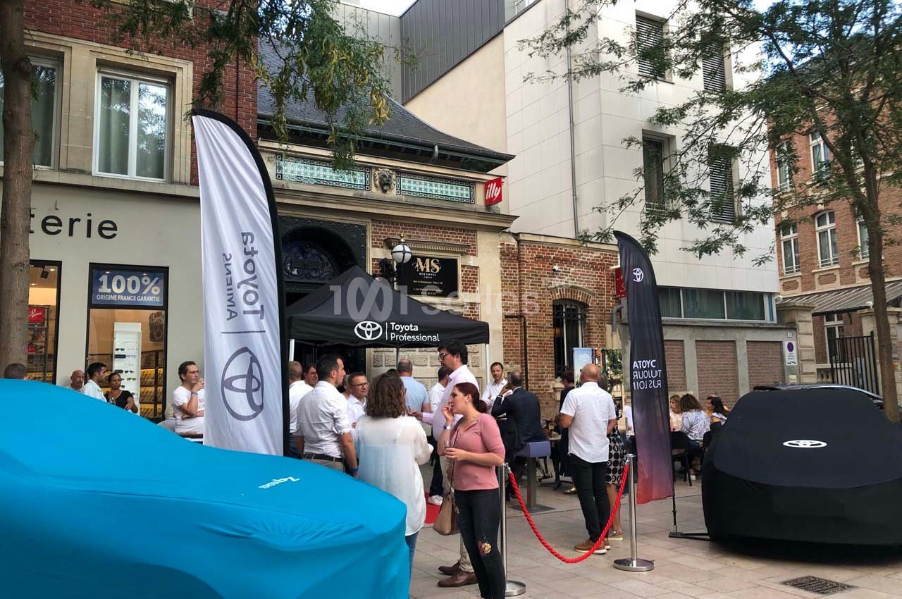 Rassemblement de personnes devant un stand Toyota avec des voitures couvertes, dans une rue bordée de bâtiments.