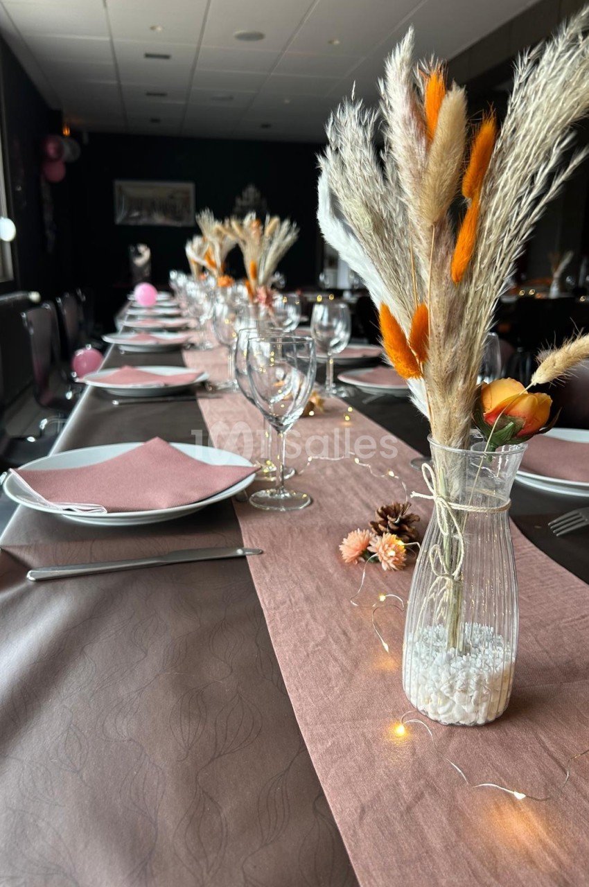 Table dressée avec une nappe rose, des assiettes, des verres et des décorations florales séchées dans des vases.