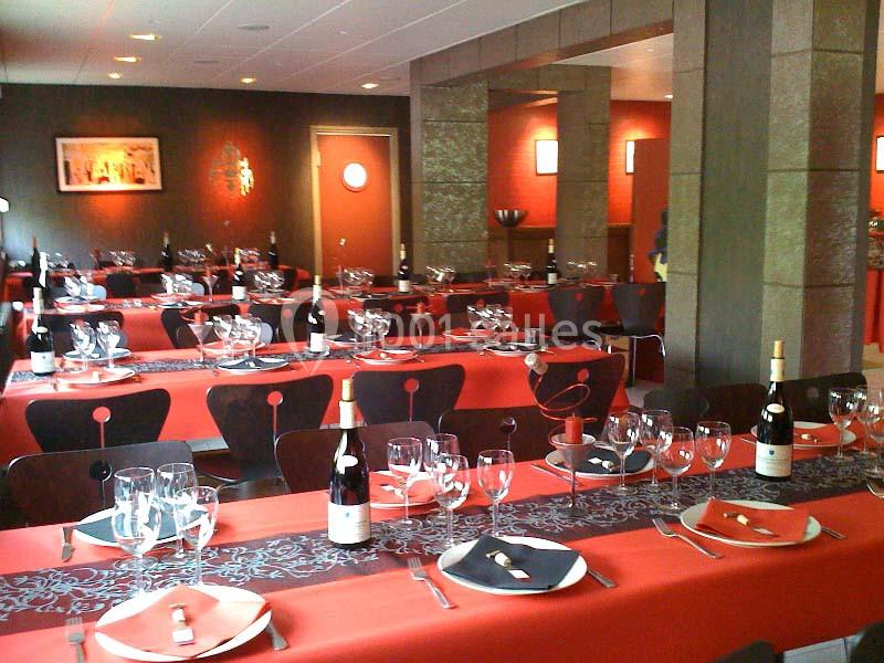 Salle de restaurant avec tables dressées, nappes rouges, verres, assiettes et bouteilles de vin.