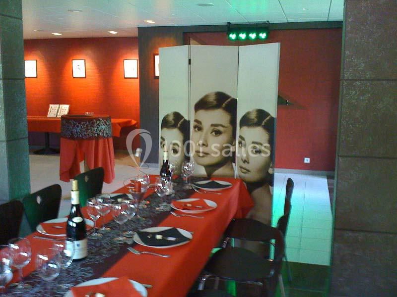 Paravent décoratif avec portraits féminins dans une salle rouge aménagée pour un repas, tables dressées avec nappes rouges.