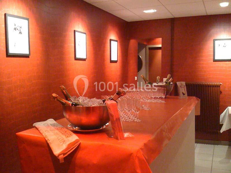 Salle avec un comptoir recouvert d'une nappe rouge, des bouteilles de champagne dans un seau et des verres alignés.