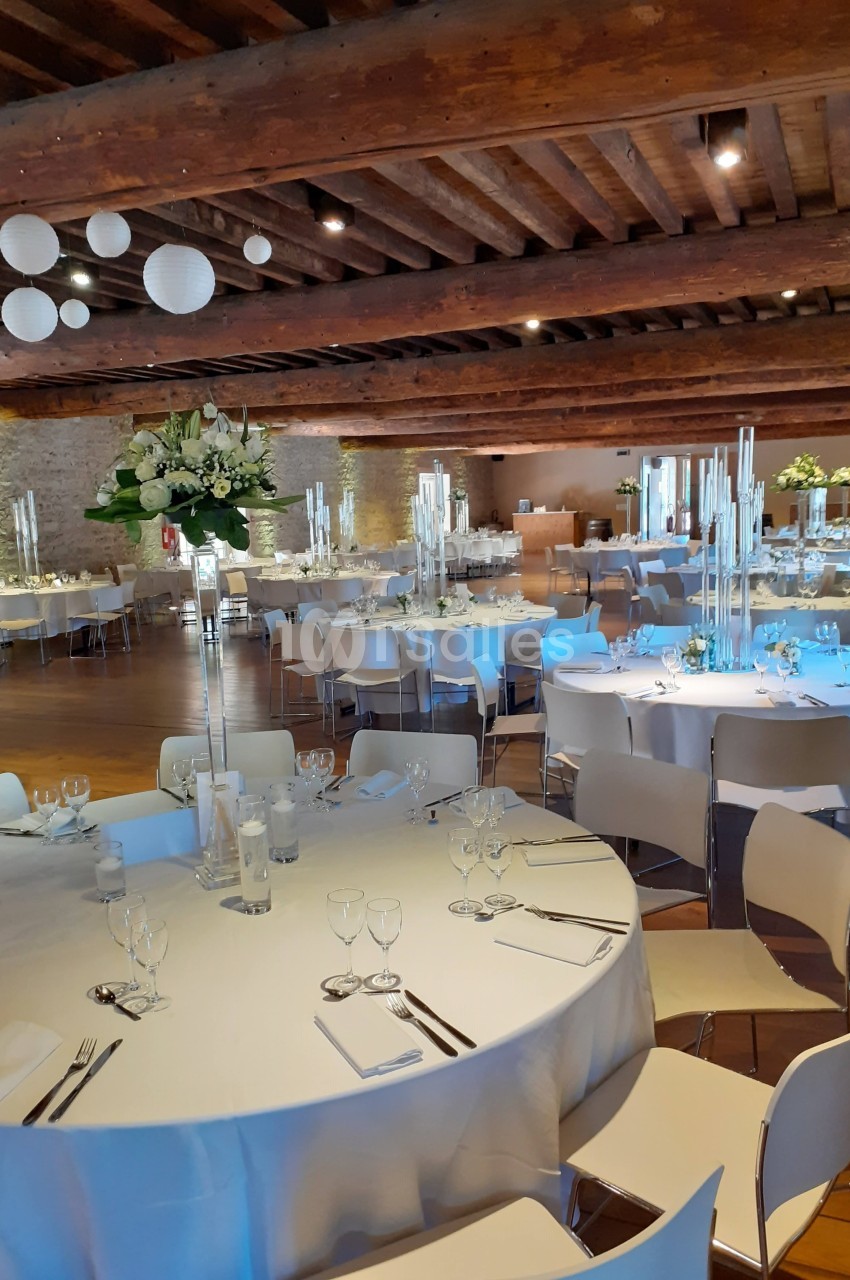 Salle de réception décorée avec des tables rondes dressées, des chandeliers et des fleurs blanches, sous un plafond en bois.