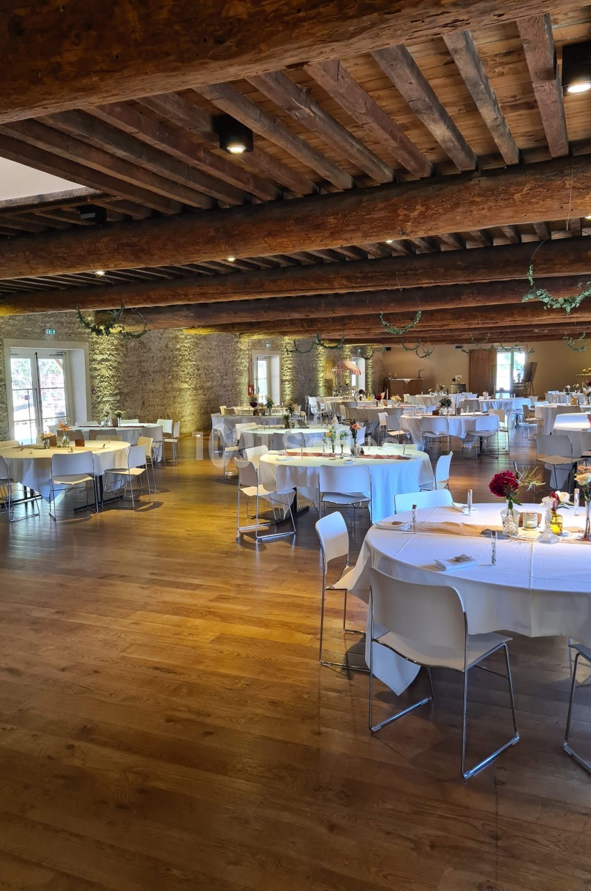 Salle de réception avec tables rondes dressées, nappes blanches, décorations florales et poutres apparentes au plafond.