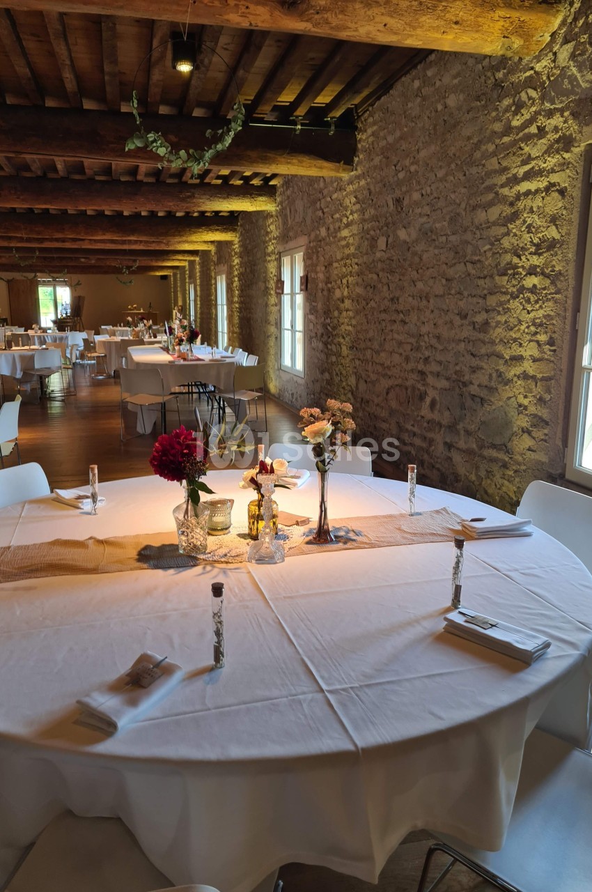 Salle de réception avec murs en pierre, tables rondes dressées avec nappes blanches et décorations florales.