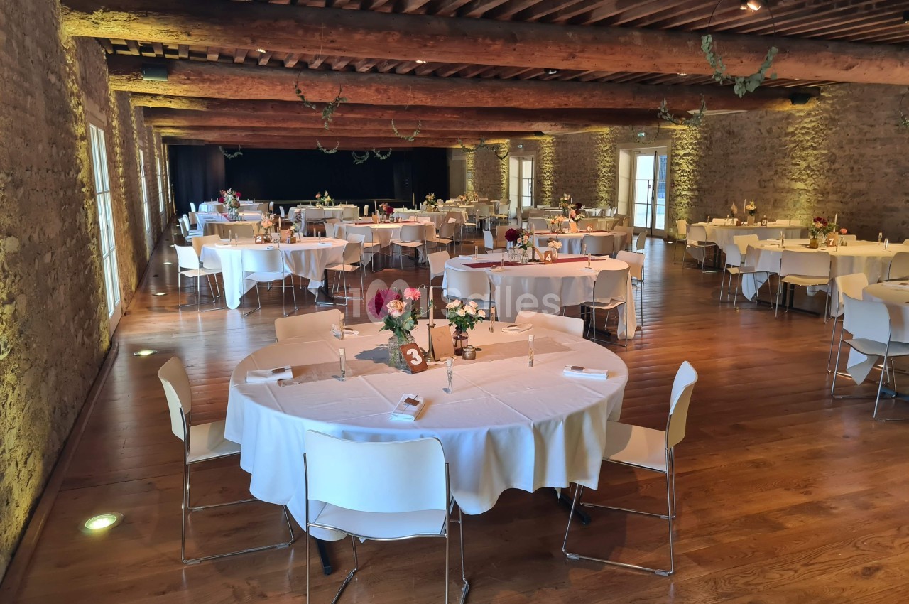 Salle de réception avec tables rondes dressées, nappes blanches, chaises modernes et poutres apparentes au plafond.