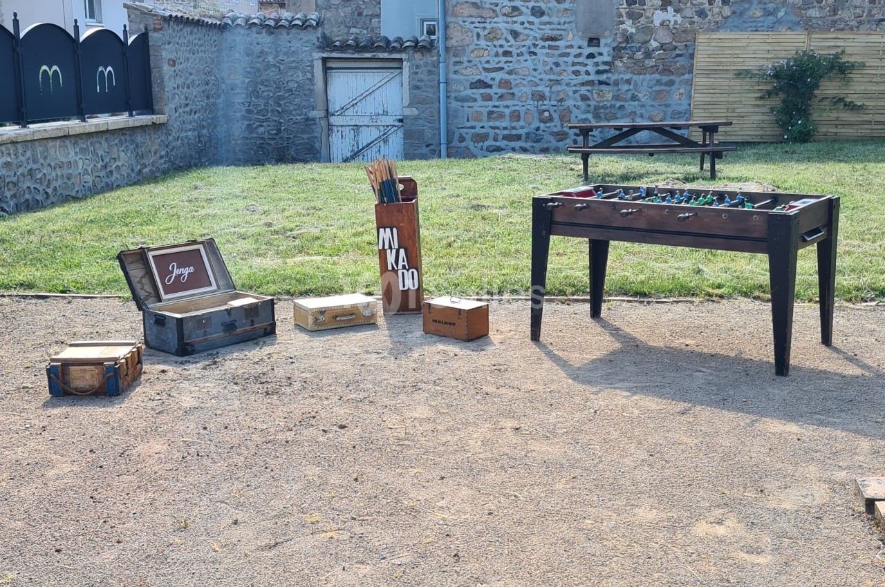 Jeu de baby-foot, valises et jeux en bois disposés en extérieur sur une surface en gravier près d'une pelouse.