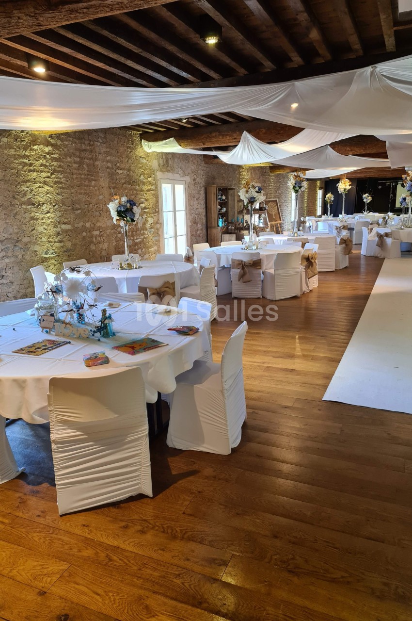 Salle de réception décorée avec des tables rondes, nappes blanches, chaises houssées et arrangements floraux.