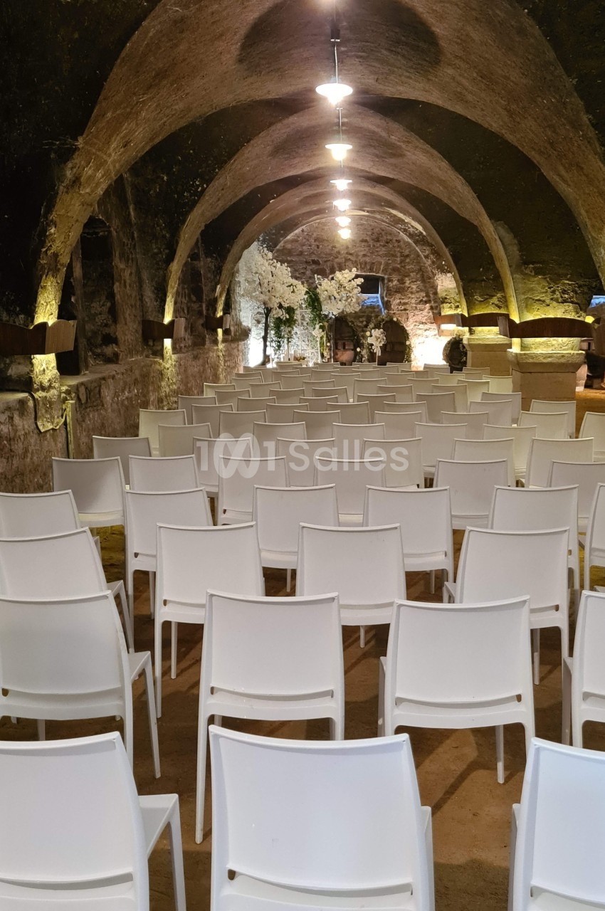 Salle voûtée en pierre avec des rangées de chaises blanches alignées face à une scène éclairée.