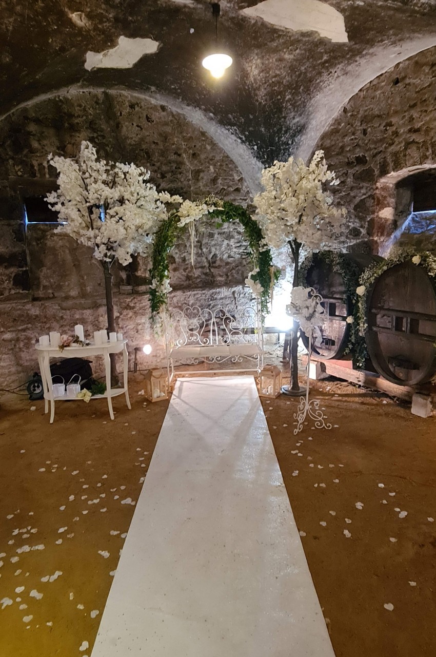 Allée blanche menant à une arche décorée de fleurs blanches et vertes dans une cave voûtée en pierre.