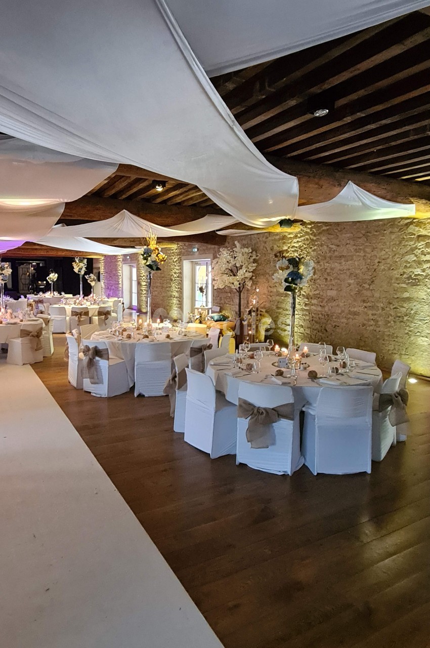 Salle de réception décorée avec des tables rondes, nappes blanches, chaises ornées de nœuds et plafond drapé.