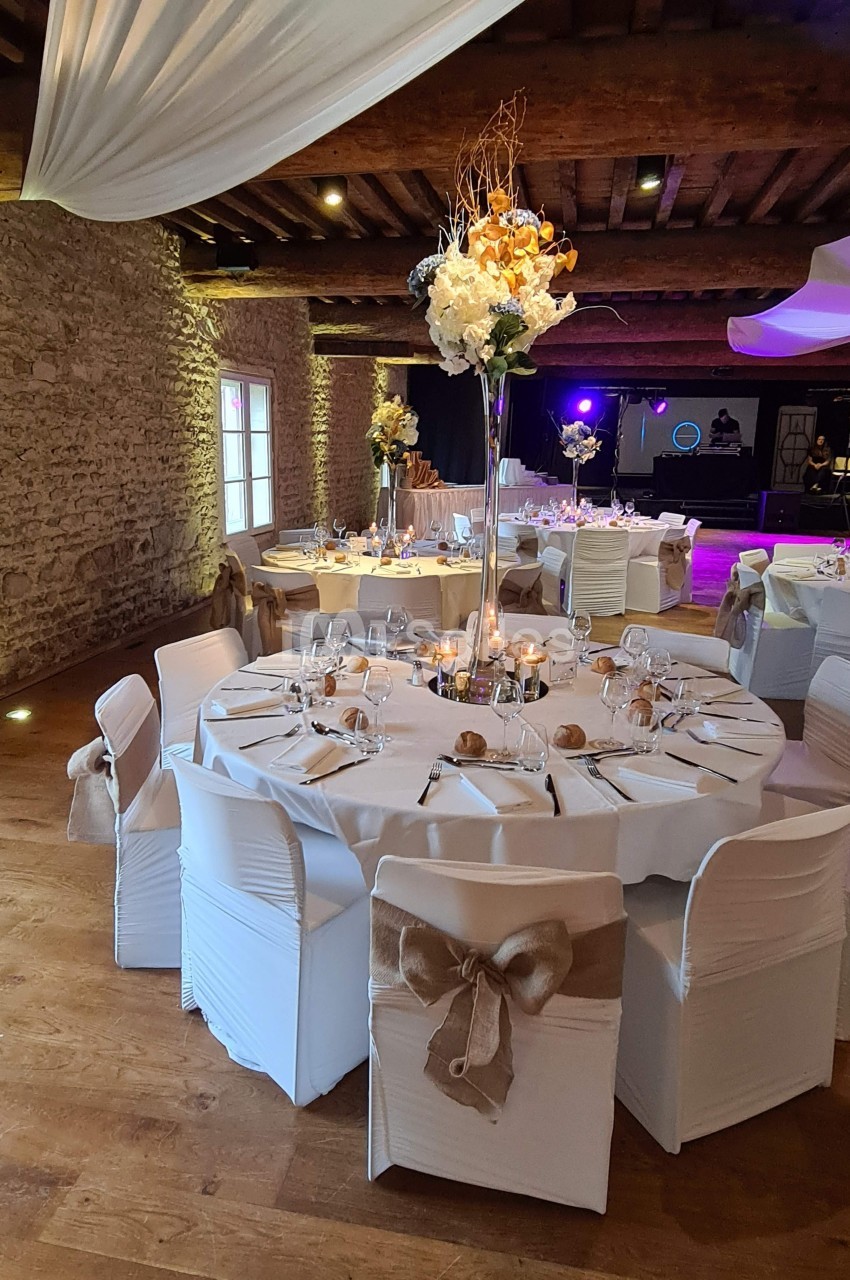 Salle de réception décorée avec des tables rondes, nappes blanches, chaises ornées de nœuds et centre de table floral.