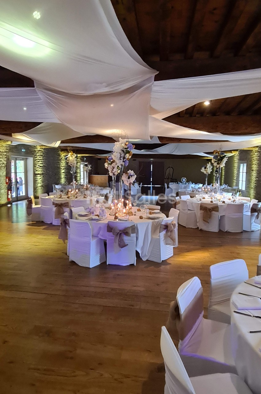 Salle de réception décorée avec des tables rondes, nappes blanches, centres de table fleuris et voilages suspendus.