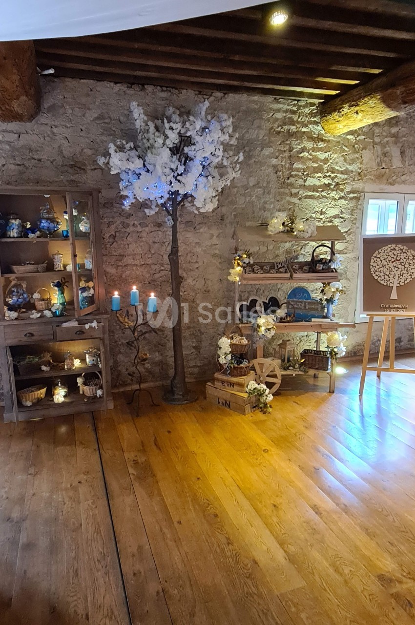 Salle en pierre avec décoration rustique, étagères illuminées, arbre artificiel fleuri et tableau sur chevalet.