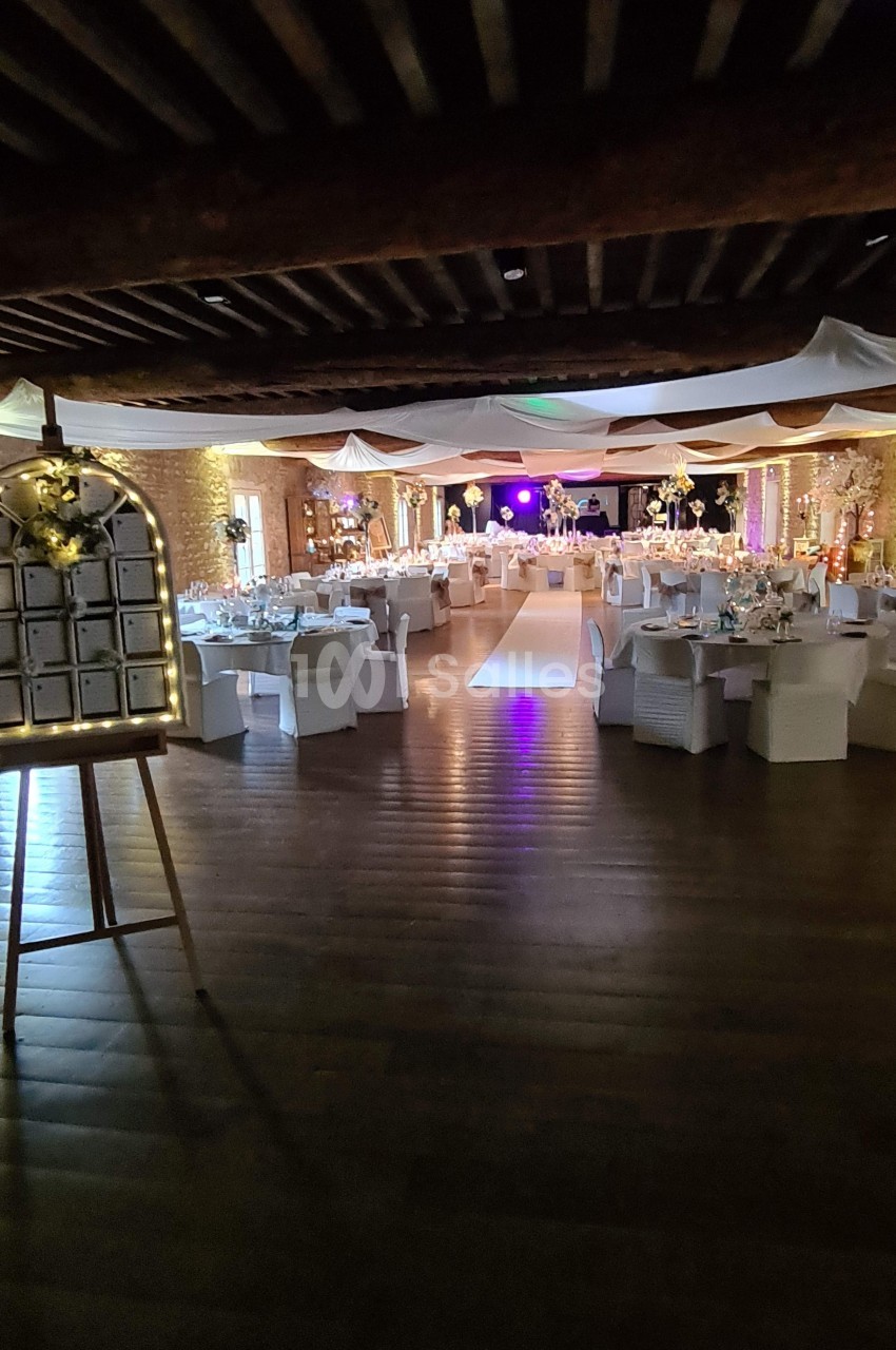 Salle de réception décorée pour un événement, avec tables dressées, éclairages chaleureux et allée centrale.