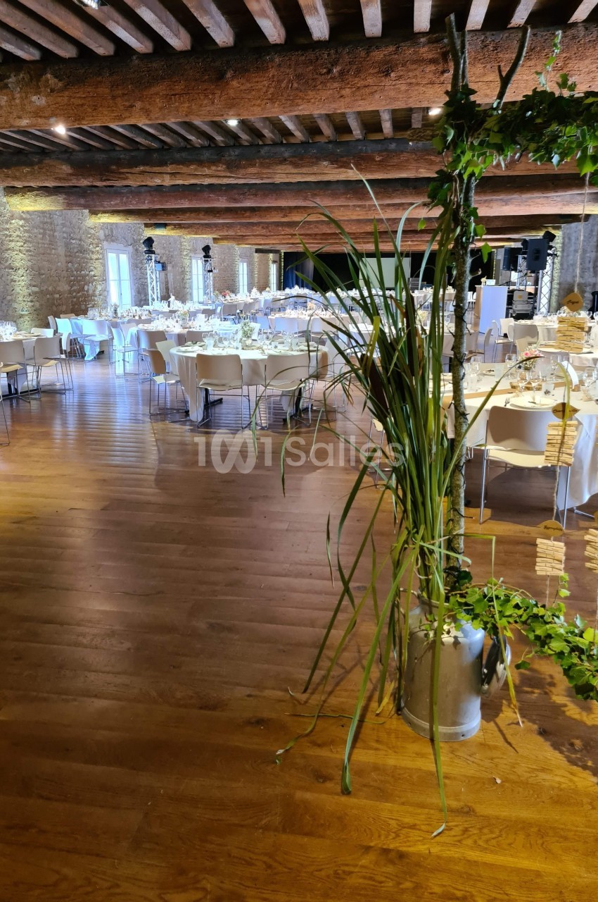 Salle de réception avec tables rondes dressées, poutres apparentes et décoration végétale au premier plan.