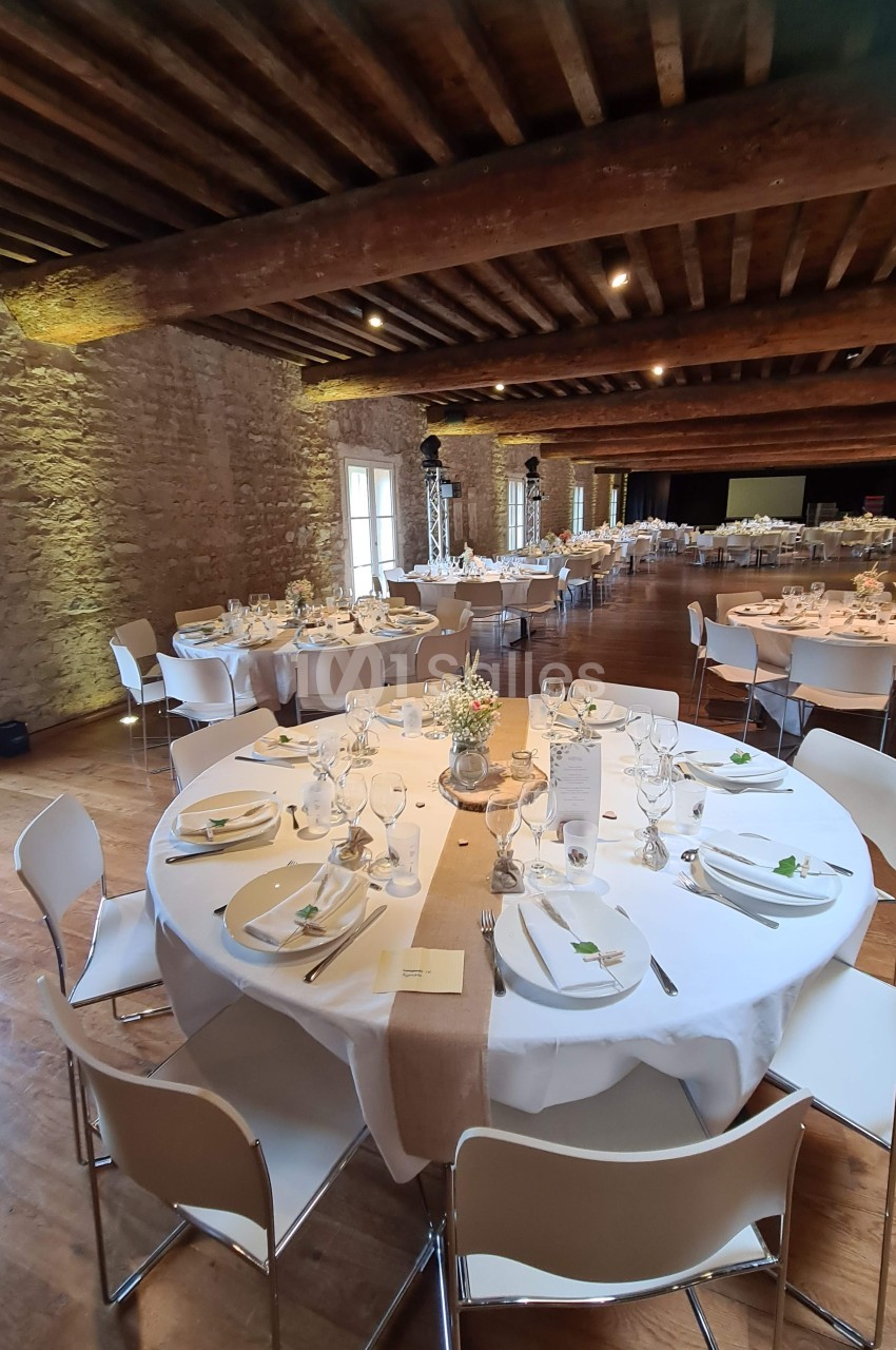 Salle de réception avec tables rondes dressées, nappes blanches, chaises modernes et murs en pierre apparente.