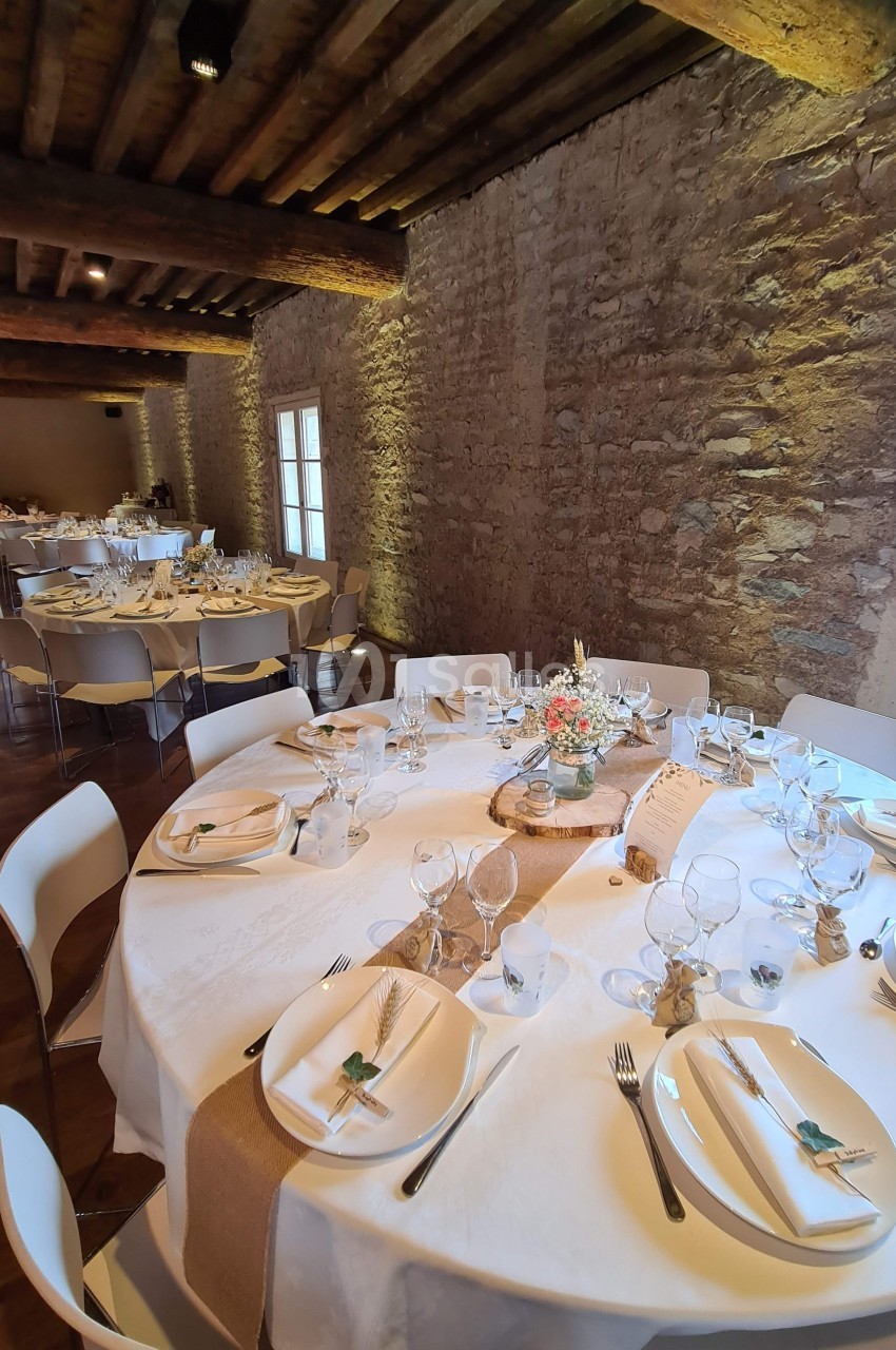 Salle de réception avec tables rondes dressées, nappes blanches, décorations florales et murs en pierre apparente.
