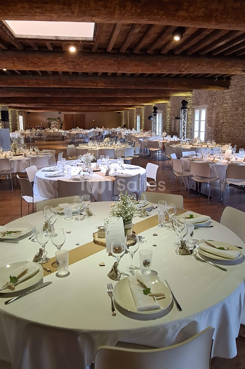 Salle de réception avec tables rondes décorées pour un événement, dans un espace aux murs en pierre et poutres apparentes.