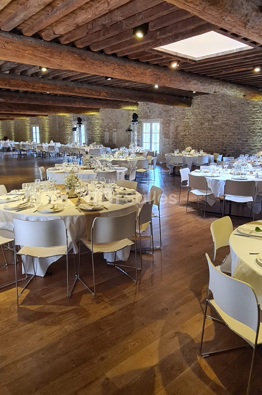 Salle de réception avec des tables rondes dressées, nappes blanches, chaises modernes et murs en pierre apparente.