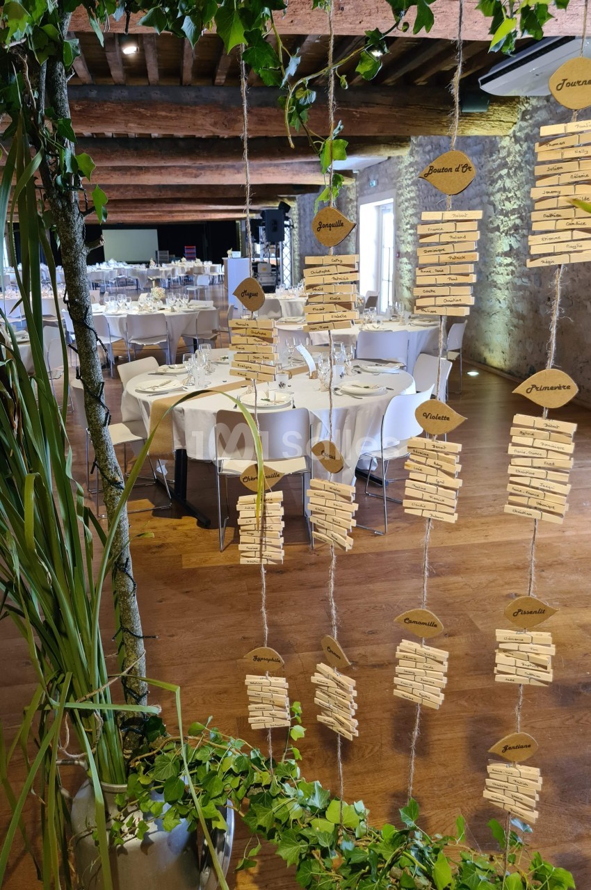 Décoration de mariage avec des marque-places suspendus en bois, tables dressées en arrière-plan dans une salle rustique.