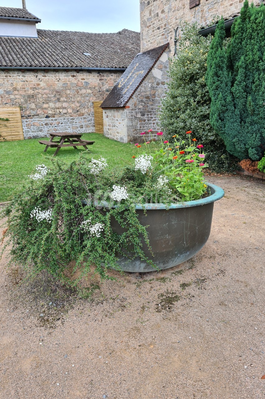 Bac rond en métal contenant des plantes fleuries, situé dans une cour avec pelouse, arbres et bâtiments en pierre.