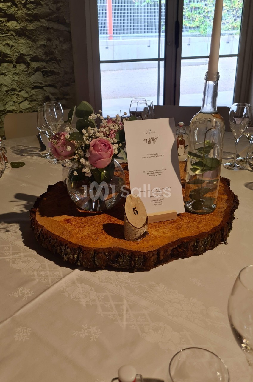 Centre de table avec rondin de bois, vase de fleurs, menu, bougie et bouteille d'eau, sur une nappe blanche.