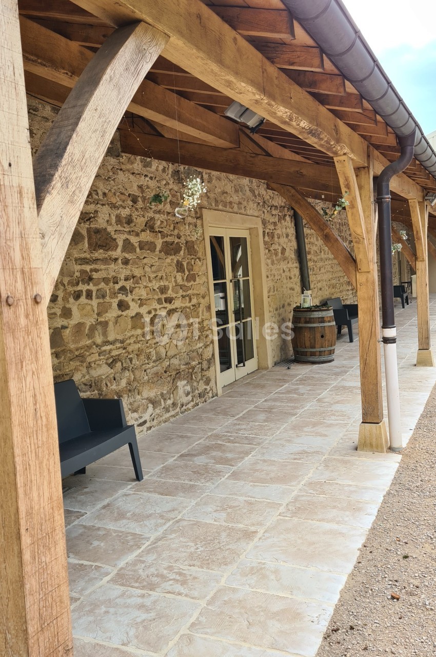 Terrasse couverte avec poutres en bois, murs en pierre, sol carrelé, bancs noirs et tonneau décoratif.
