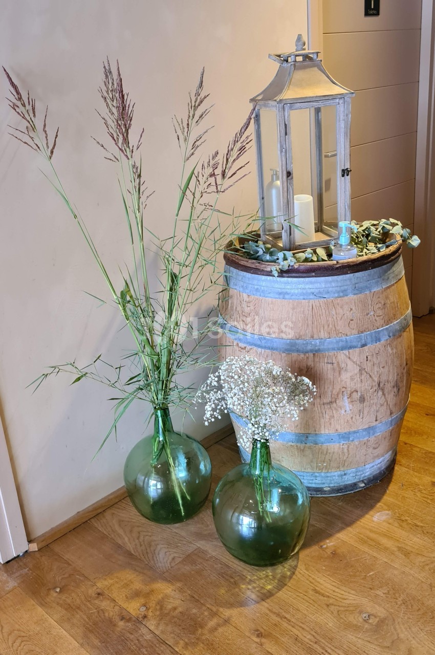Deux vases en verre vert avec des fleurs séchées posés au sol près d'un tonneau décoré d'une lanterne et de bougies.